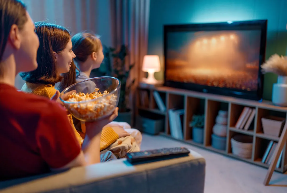 Tre persone sul divano guardano la TV con popcorn. Lo schermo mostra un concerto. Sotto la TV, una libreria con libri e oggetti decorativi in una stanza buia.