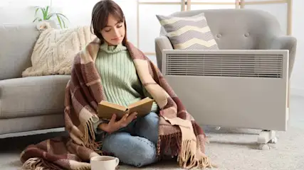 Frau sitzt am Boden mit einer Decken über den schultern und liest ein Buch, daneben steht ein Heizgerät