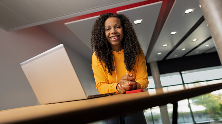 Une femme souriante en pull jaune près d'un ordinateur portable sur une table en bois, avec un plafond moderne en arrière-plan.