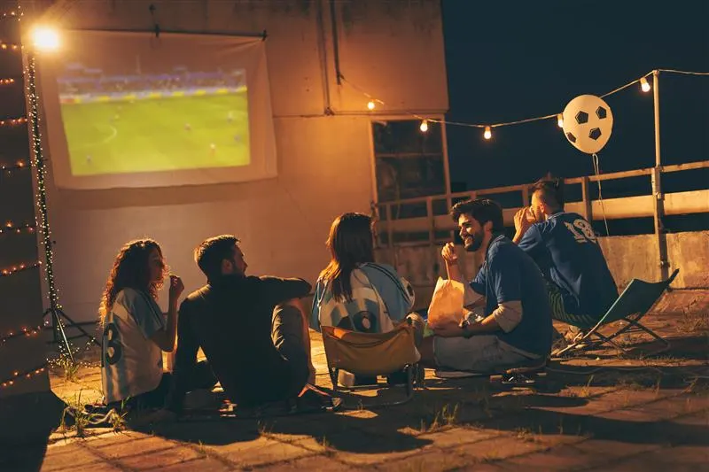 Cinq personnes regardent un match de football projeté sur un écran extérieur la nuit, sous guirlandes lumineuses. Un ballon blanc flotte.