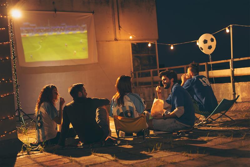 Cinque persone, alcune con maglie sportive, guardano una partita di calcio proiettata di notte su uno schermo esterno, illuminate da luci e con un pallone.