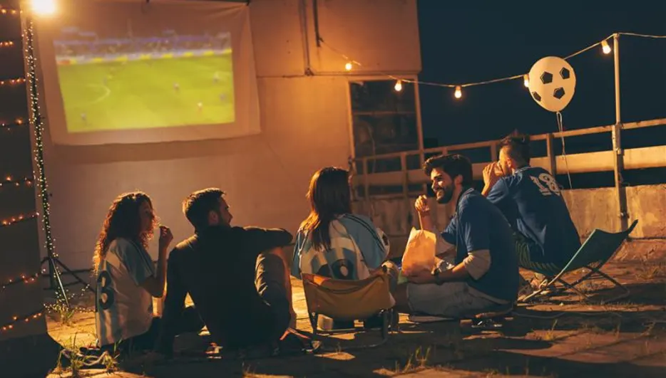 Fünf Personen sehen nachts draußen ein Fußballspiel auf einer weißen Leinwand. Lichterketten und ein Fußballballon schmücken den Hintergrund.