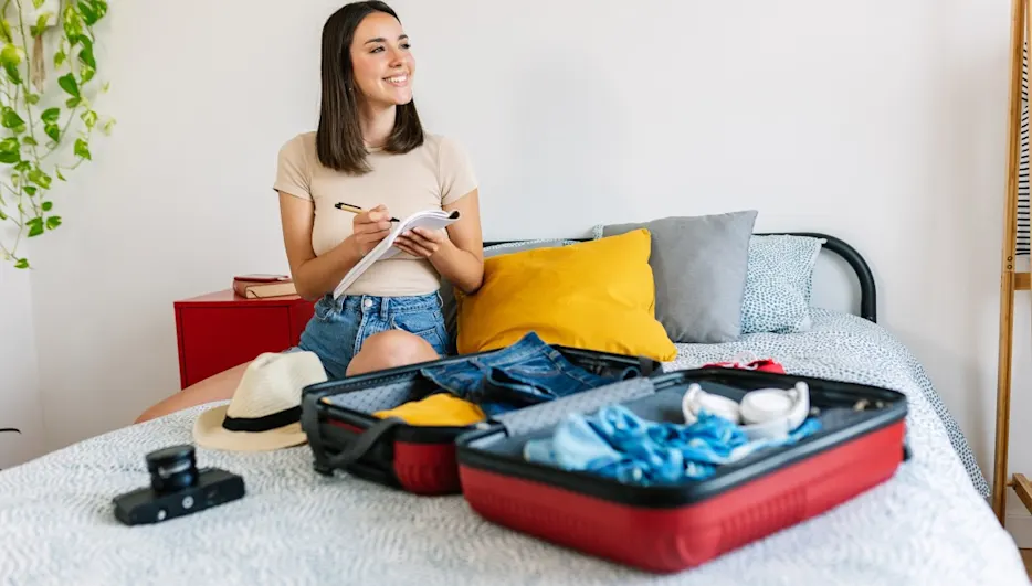 Junge Frau sitzt auf dem Bett neben einem halbgepackten Koffer und arbeitet ihre Packliste durch.