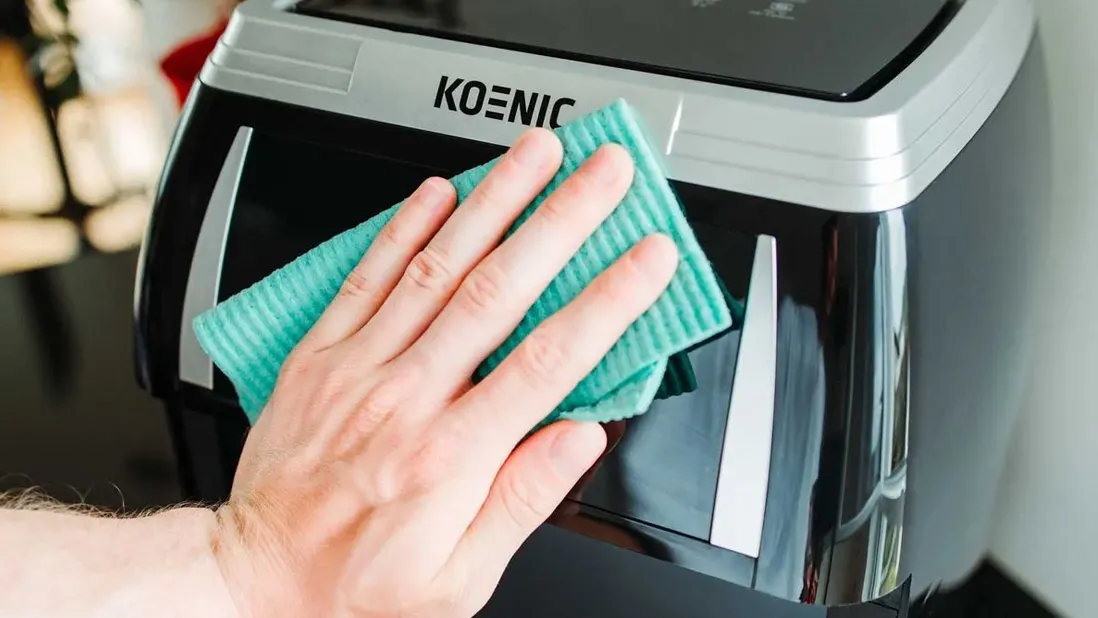 Une main essuie un appareil de cuisine noir et argenté de marque KOENIC avec un chiffon de nettoyage turquoise.
