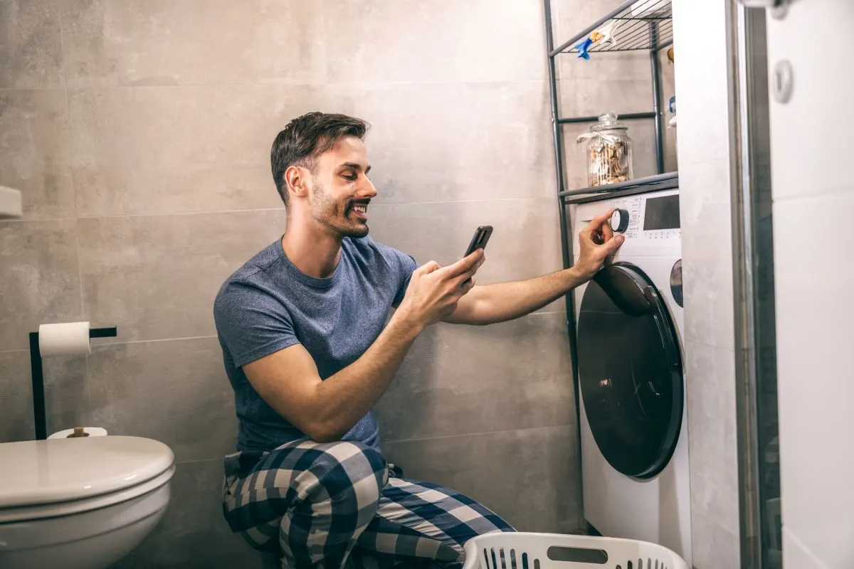 Un uomo in bagno davanti a una lavatrice, tiene in mano un cellulare e gira una manopola sulla lavatrice.
