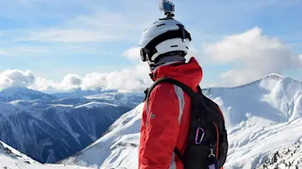 Person mit Helm und Rucksack trägt eine Action Cam in den Bergen im Schnee.
