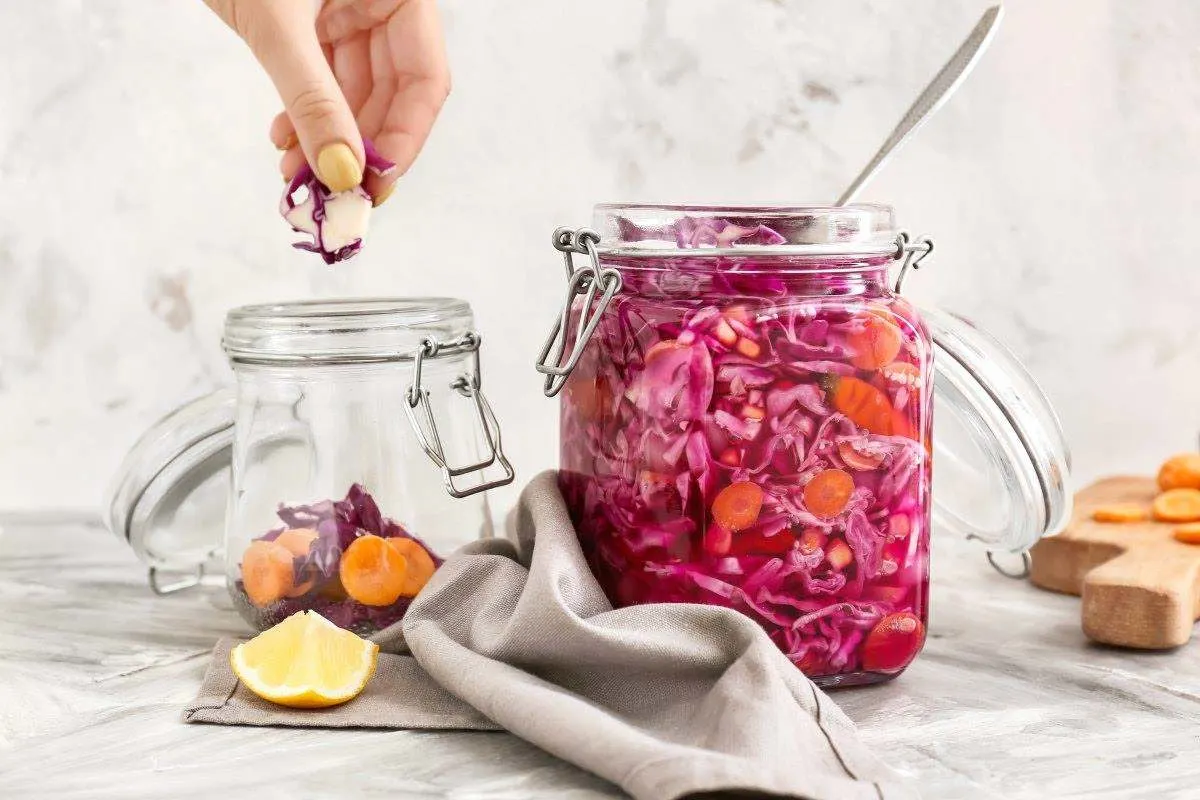 Fermentierter Rotkohl und Karotten in einem grossen Einmachglas, während eine Hand weitere Kohlstücke hinzugibt.