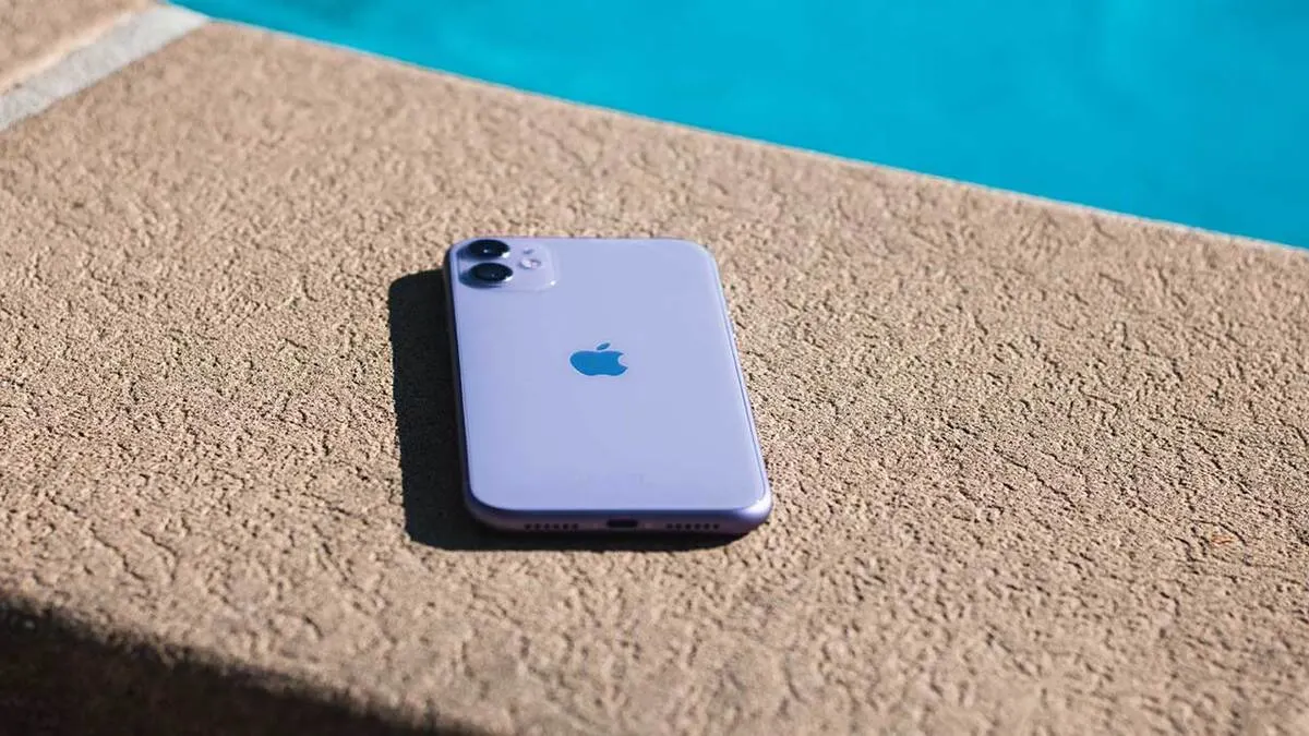 Un telefono viola con logo a forma di mela è appoggiato su una superficie beige ruvida, con acqua turchese sullo sfondo.