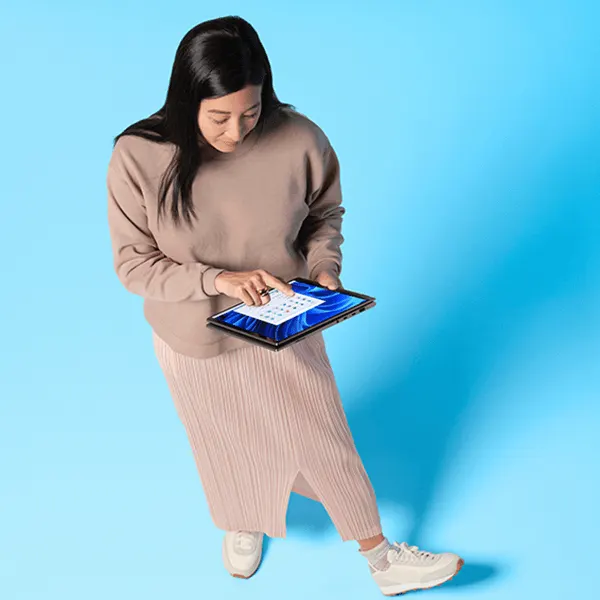 Une femme vêtue d'un pull et d'une jupe beiges tient un Windows Convertible et sélectionne une application via l'écran tactile.