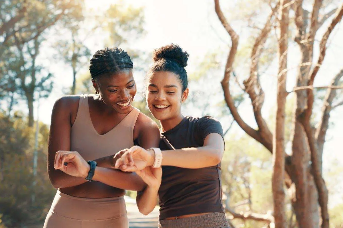 Zwei lachende Frauen in Sportkleidung vergleichen beim Laufen im Wald die Anzeigen ihrer Fitness-Tracker am Handgelenk.