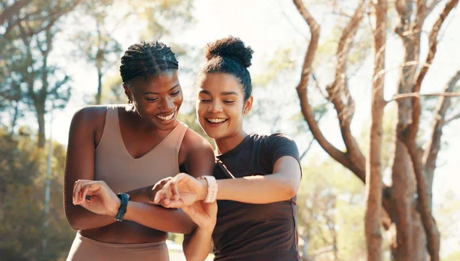 Deux femmes riantes en tenue de sport comparent, en courant en forêt, les données de leur bracelet fitness au poignet.