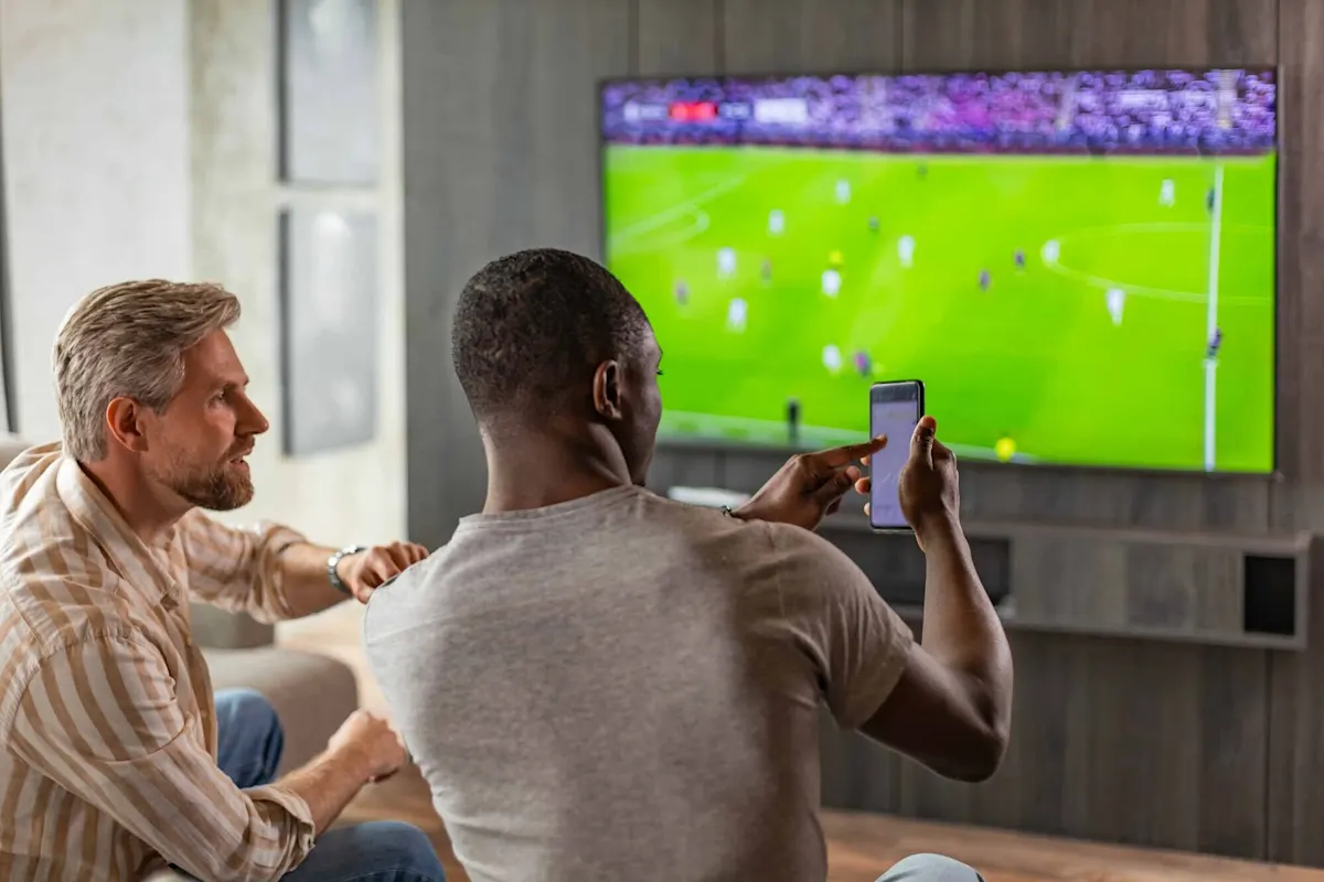 Zwei Männer sitzen im Wohnzimmer vor dem Fernseher mit laufendem Fussballmatch. Einer zeigt dem anderen etwas auf dem Smartphone.