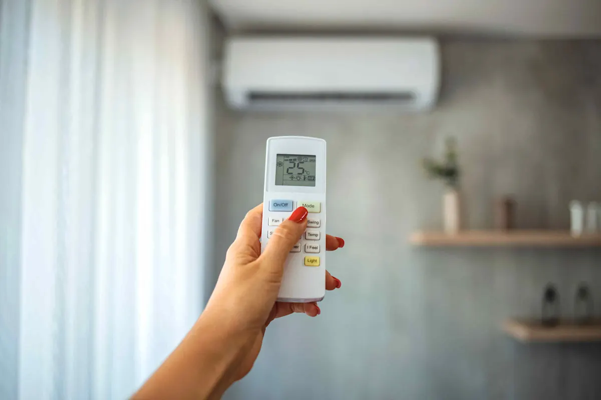 Une main tient une télécommande de climatiseur avec un affichage de température de 25°C au premier plan, à l'arrière-plan, le climatiseur est visible sur le mur.