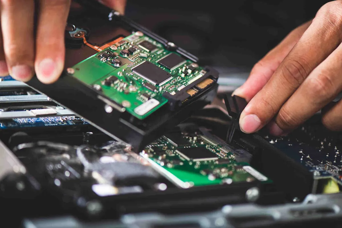 Due mani riparano un computer aperto; sui circuiti stampati verdi sono visibili chip e connettori. 