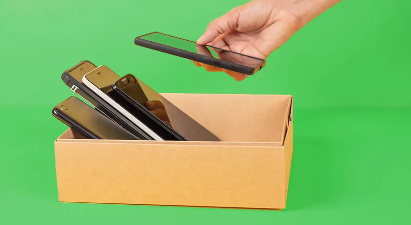 Una mano tiene in mano un telefono cellulare nero sopra una scatola piena di telefoni cellulari, su uno sfondo verde.