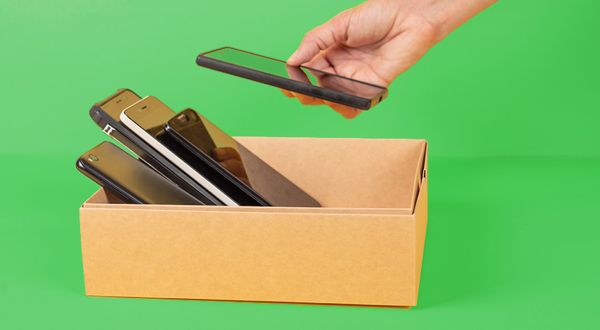 Une main tient un téléphone portable noir au-dessus d’un carton rempli de téléphones portables, sur un fond vert.