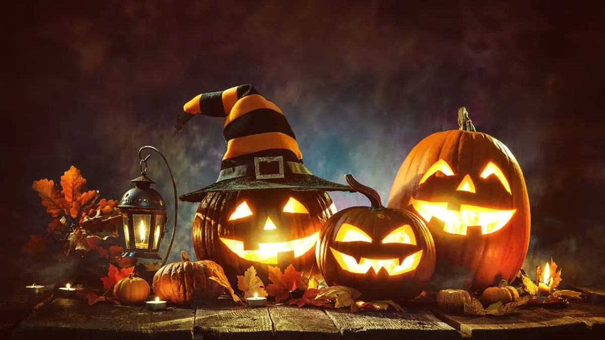 Tre zucche di Halloween intagliate e sorridenti, di cui una con un cappello da strega a strisce arancioni e nere, circondate da decorazioni autunnali come candele, lanterne, foglie e zucche decorative.