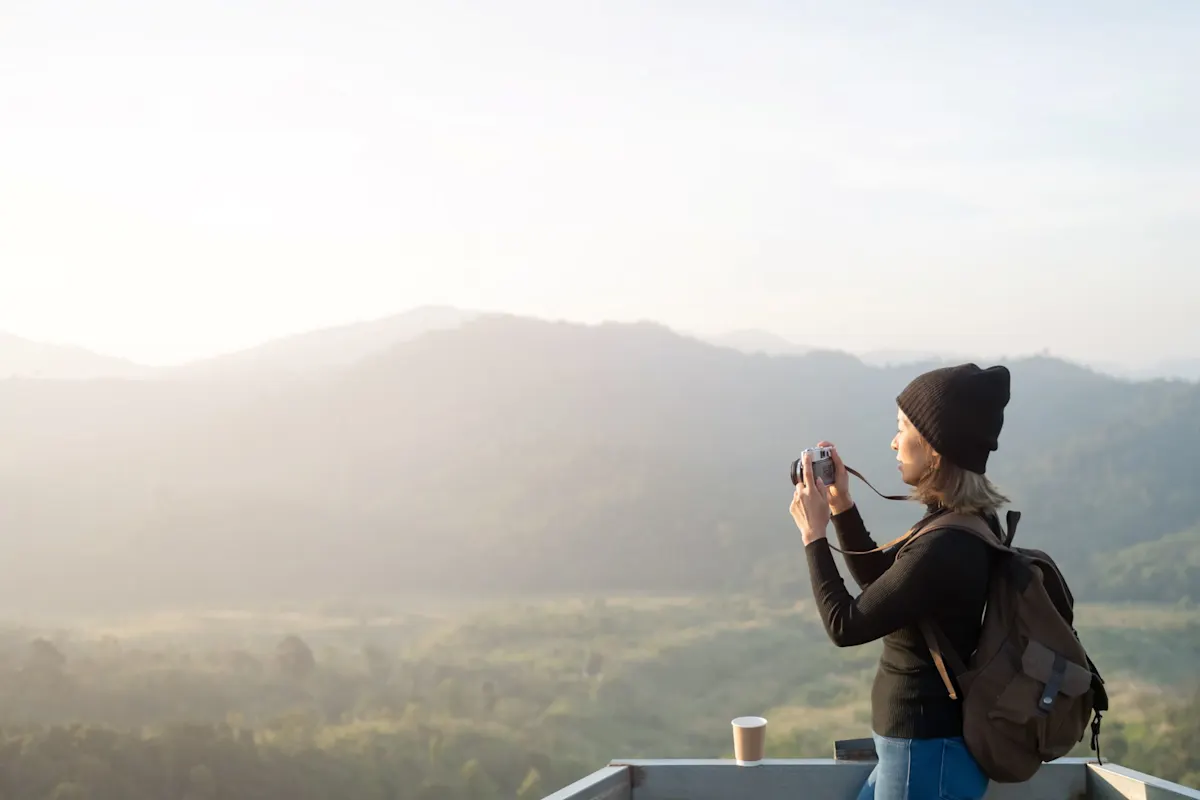 Une femme avec un bonnet et un sac à dos photographie un vaste paysage de montagne dans la lumière matinale avec un appareil photo compact.