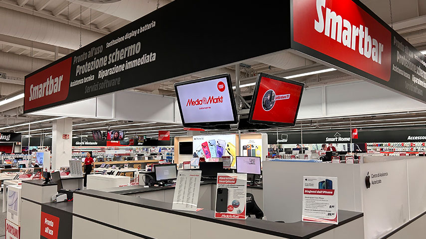 Interno di un negozio di elettronica con scaffali di prodotti, insegne "Smartbar" e schermi sospesi. Banchi vendita visibili in primo piano.