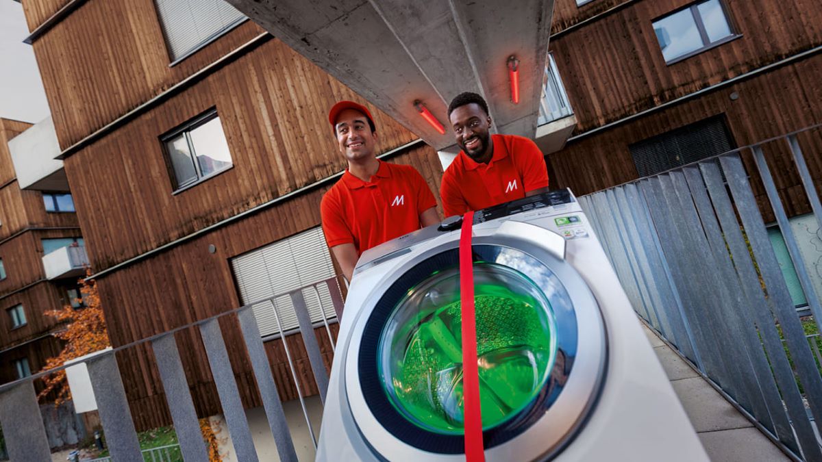 Zwei MediaMarkt-Mitarbeiter in roter Arbeitskleidung liefern eine AEG Waschmaschine aus.