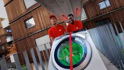 Zwei MediaMarkt-Mitarbeiter in roter Arbeitskleidung liefern eine AEG Waschmaschine aus.