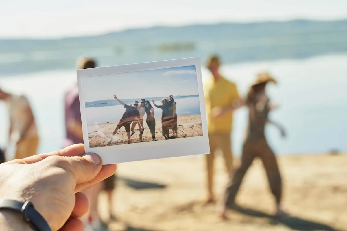 Une main tient une photo Polaroid fraîchement prise devant une scène de plage avec plusieurs personnes.