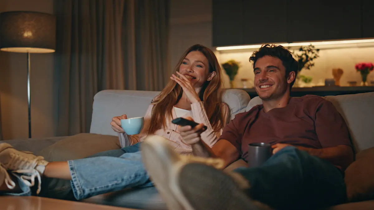 Due persone sono sedute comodamente su un divano, bevono da tazze e guardano sorridendo la televisione insieme.