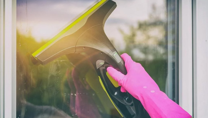 Eine Hand mit einem pinkfarbenen Gummihandschuh hält einen grauen Fensterreiniger mit gelben Akzenten an einer Fensterscheibe.