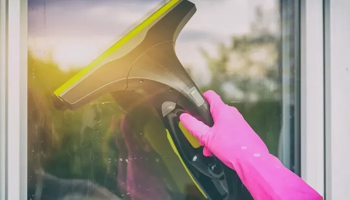 Une main gantée en rose nettoie une vitre avec un outil de nettoyage jaune et noir, devant un cadre de fenêtre blanc.