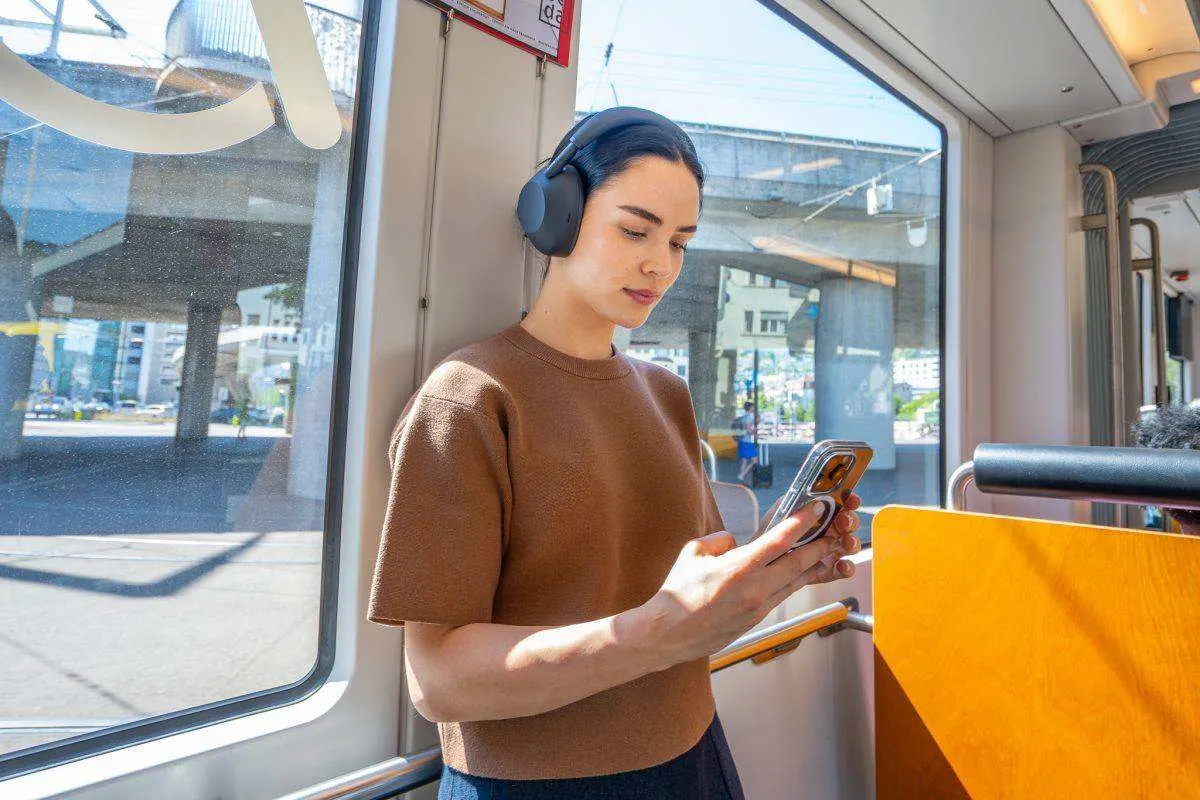 Donna che indossa le cuffie over-ear Sony WH-1000XM6 in nero, mentre guarda il proprio smartphone su un tram. 