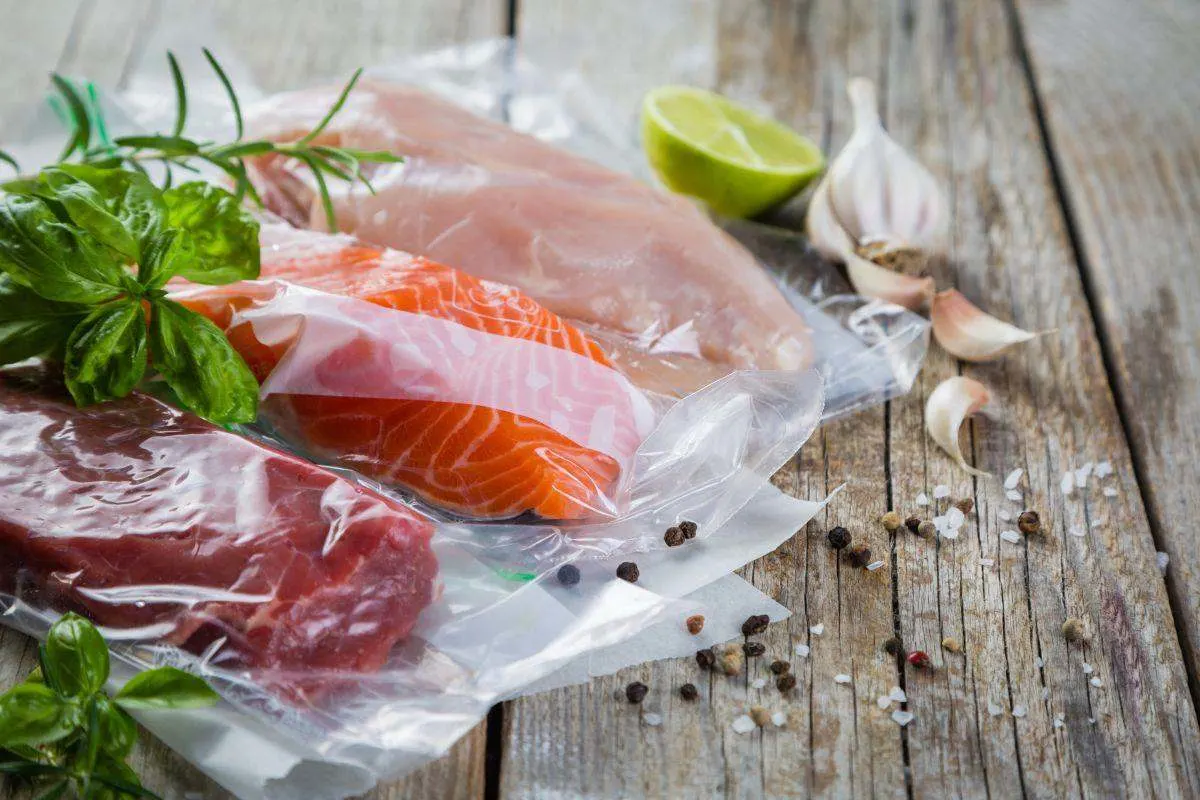 Vakuumverpackte Stücke von Lachs, Fleisch und Geflügel auf einem Holztisch, dekoriert mit Kräutern, Limette, Knoblauch und Gewürzen.