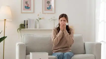 Frau sitzt auf der Couch, hält sich ein Taschentuch vor das Gesicht, im Vordergrund steht ein Luftreiniger.