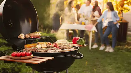 Barbecue boule rempli de viande et de légumes au premier plan, tandis qu’à l’arrière-plan plusieurs personnes sont assises ensemble dans le jardin.