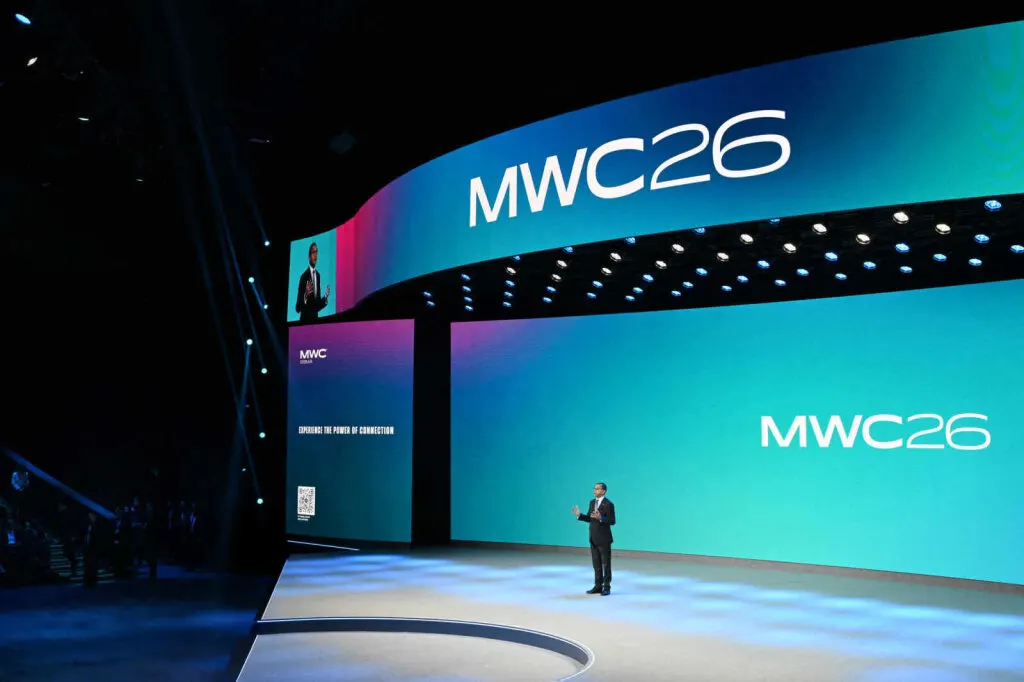Moderatore su un palco al MWC; dietro di lui sulla parete compare due volte la scritta «MWC26». 