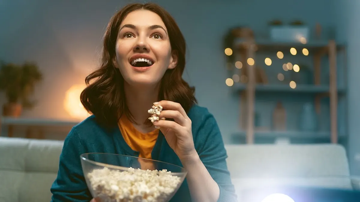 Eine Frau isst Popcorn aus einer Glasschüssel. Im Hintergrund sind ein Regal und Lichterketten zu sehen.