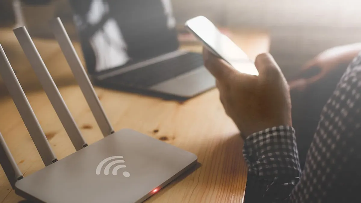 Auf einem Tisch steht ein Router mit Antennen, daneben ein Laptop und eine Person, die ein Mobiltelefon hält.