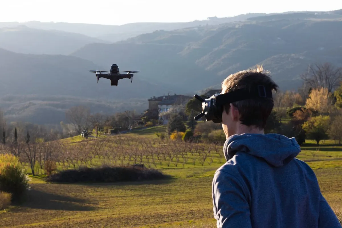 Person mit FPV-Brille steuert eine Drohne, die vor einer hügeligen Landschaft mit Feldern und Bäumen fliegt.