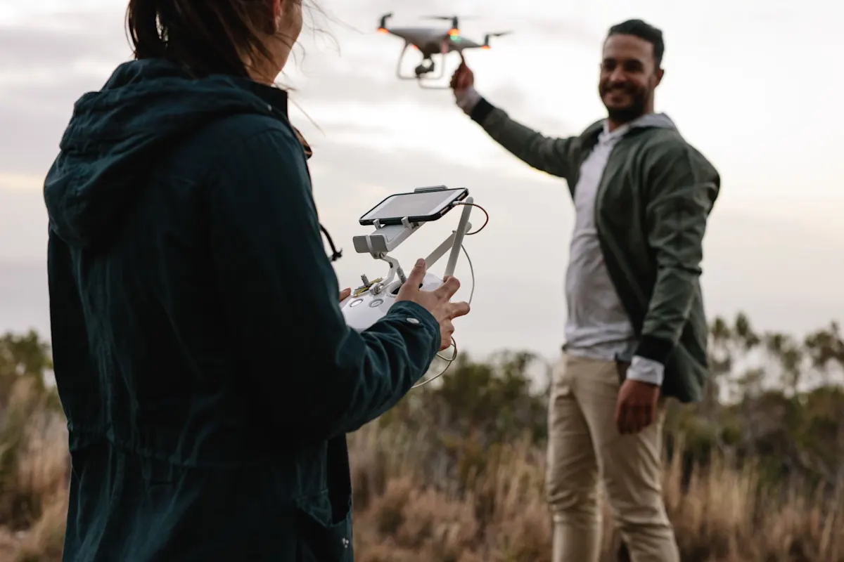Personne avec radiocommande et tablette au premier plan; une autre lance ou attrape un drone éclairé au coucher du soleil, en extérieur.