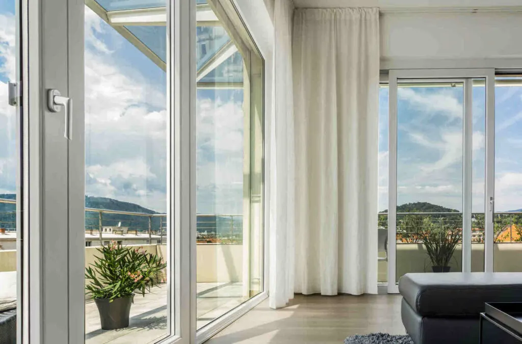 Salon lumineux avec de grandes fenêtres, des rideaux blancs et vue sur le balcon, les collines et le ciel.