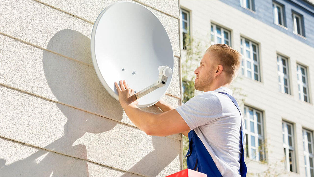 Un homme installe une antenne parabolique blanche sur un mur. Un immeuble blanc est visible en arrière-plan.