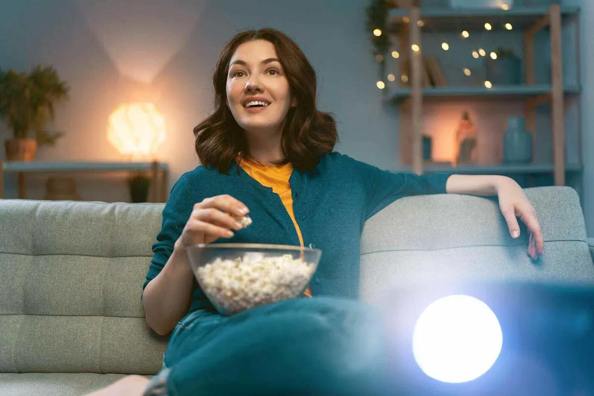 Frau sitzt mit einer Schüssel Popcorn auf einem Sofa, im Vordergrund ist ein helles Licht eines Mini Beamers zu sehen.