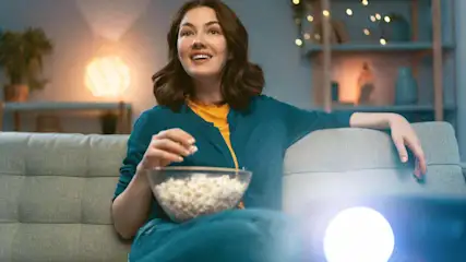 Frau sitzt mit einer Schüssel Popcorn auf einem Sofa, im Vordergrund ist ein helles Licht eines Mini Beamers zu sehen.
