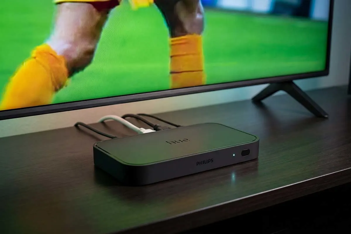 Philips Hue Sync Box liegt vor einem Fernseher, auf dem ein Fussballspiel läuft; mehrere Kabel sind angeschlossen und ein kleines Statuslicht leuchtet.