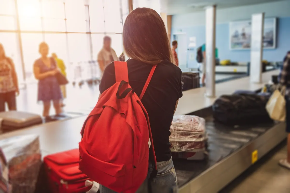 Frau mit rotem Rucksack steht am Gepäckband im Flughafen und wartet auf ihr Reisegepäck.