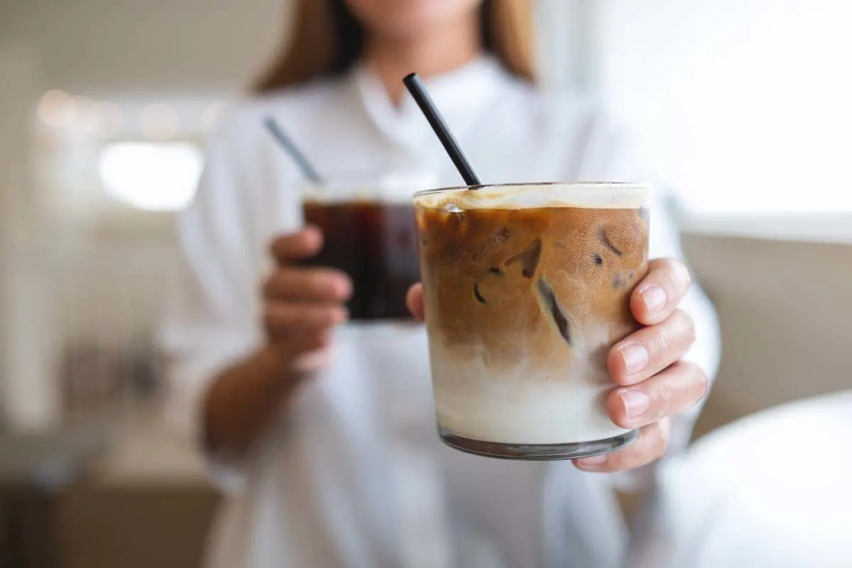 Una persona mostra alla fotocamera un bicchiere di caffè freddo con latte e cannuccia nera. 