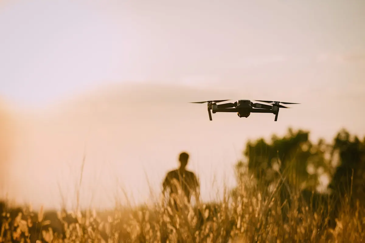 Drone volant au-dessus d’un champ au coucher du soleil, silhouette floue d’une personne à l’arrière-plan.