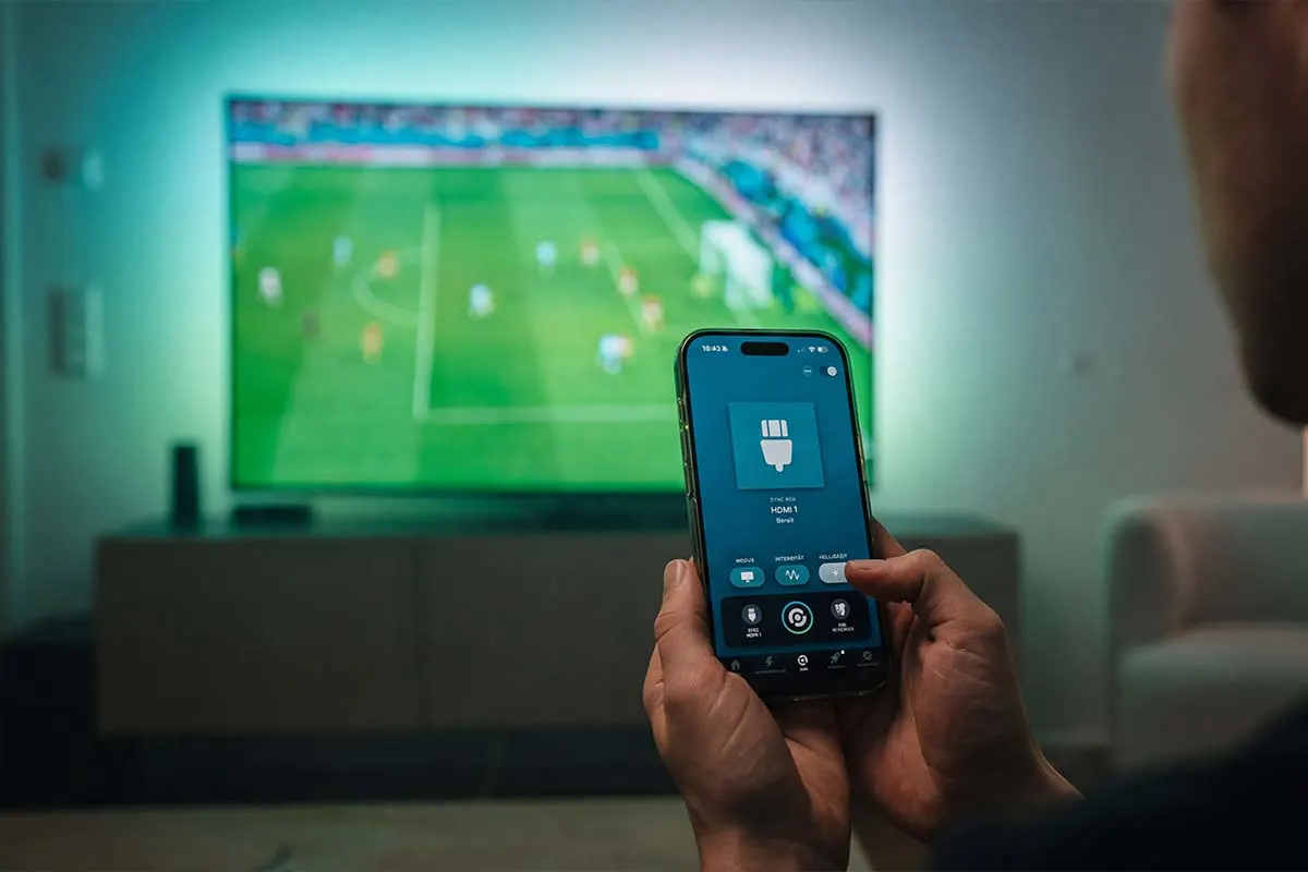Une personne contrôle la Philips Hue Sync Box à l'aide de son smartphone, tandis qu'un match de football est diffusé en arrière-plan à la télévision et qu'un éclairage d'ambiance coloré illumine le mur.