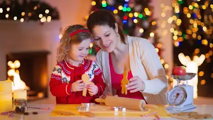 Femme avec enfant préparant des biscuits de Noël, la femme montre à l'enfant un bonhomme en pain d'épices, à droite de l'image se trouve une balance, à l'arrière-plan, on aperçoit floues les lumières de Noël.