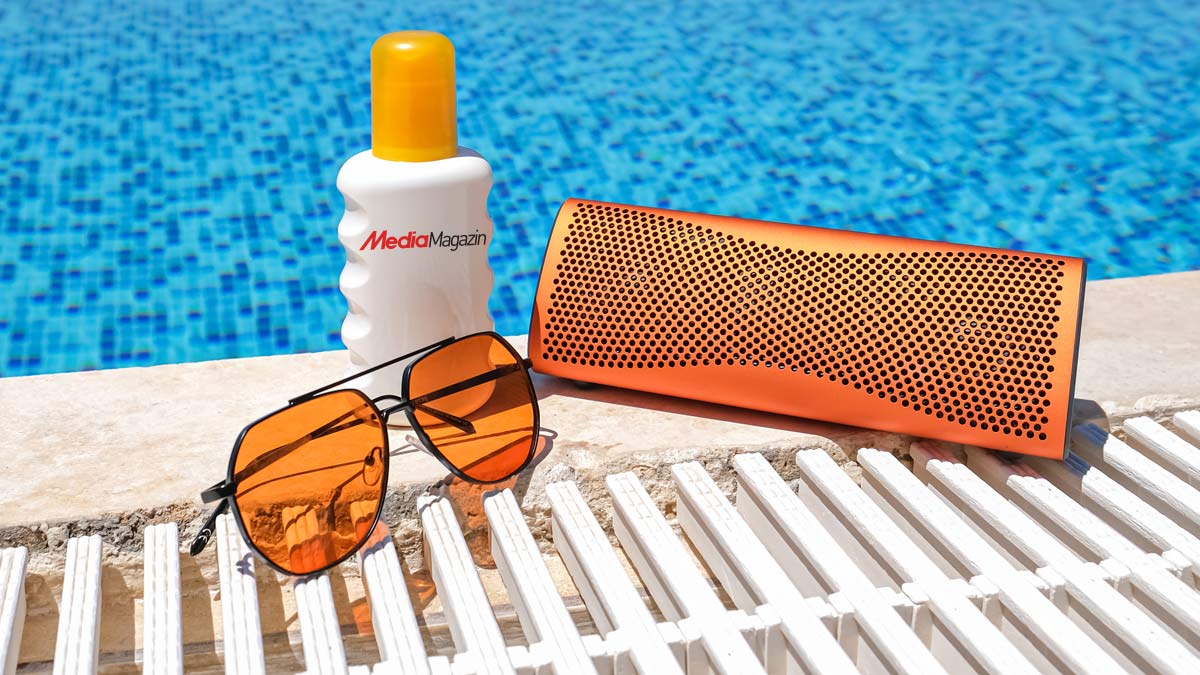 Sonnencreme, orangefarbener Lautsprecher und Brille liegen am Poolrand auf einem weissen Lattenrost.