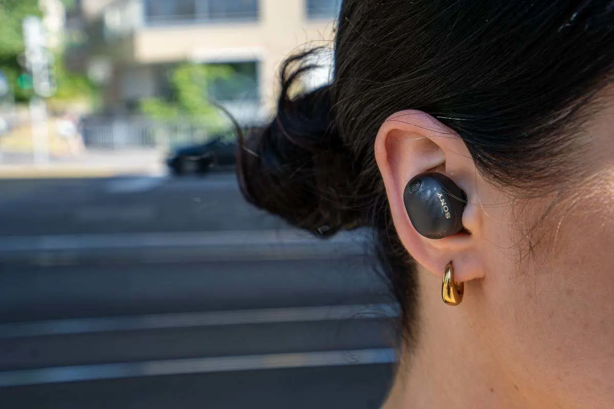 Gros plan d'un écouteur in-ear Sony WF-1000XM6 en noir porté par une femme, avec une rue urbaine en arrière-plan. 